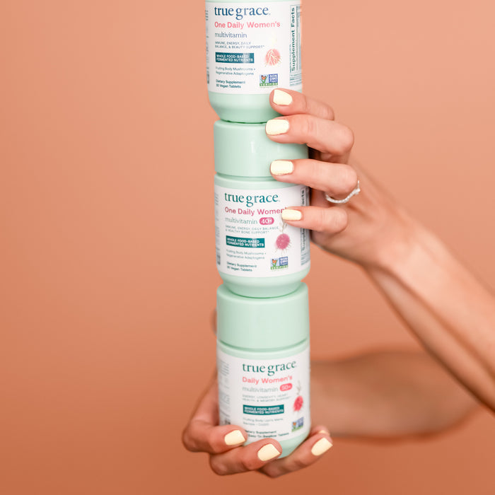 Against a peach background, a person with manicured nails holds three mint green bottles of True Grace One Daily Womens Multivitamin, enriched with Coenzyme B Vitamins. The bottles feature a pink flower design and non-GMO labeling.