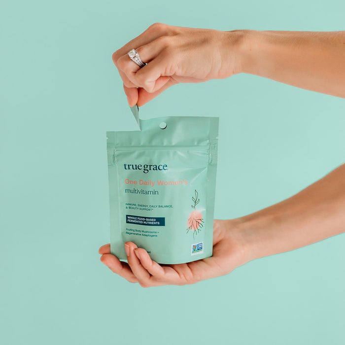 Close-up of hands holding a teal package of True Grace One Daily Womens Multivitamin Refill against a matching background, opening the resealable pack adorned with floral graphics and details, and including Coenzyme B Vitamins for enhanced wellness.