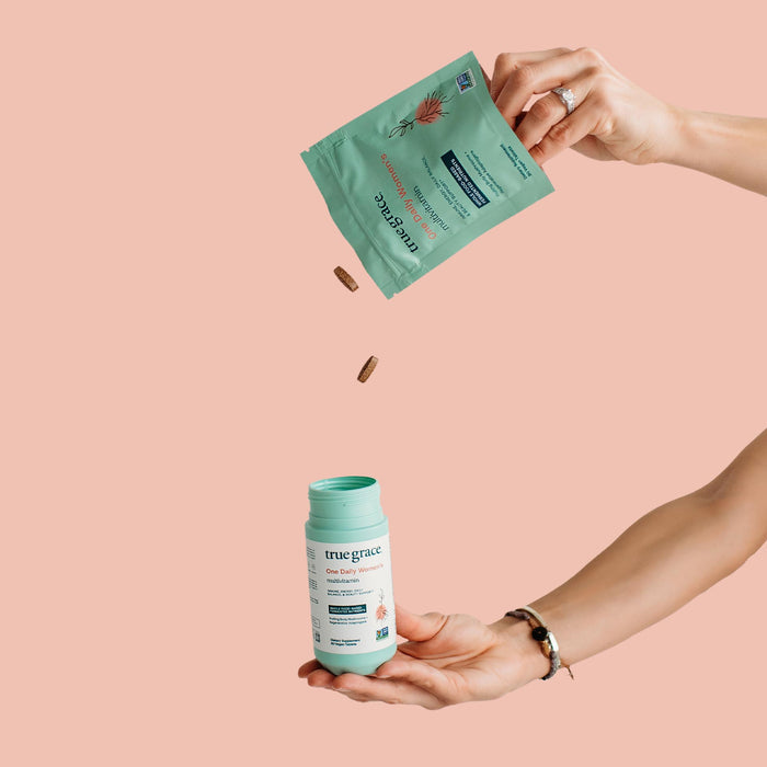 In front of a peach background, a pair of hands elegantly handle wellness items. One pours capsules from a green packet into a matching bottle, both featuring True Grace and aloe designs. The product is One Daily Womens Multivitamin Refill, using an organic mushroom blend for natural health enhancement.