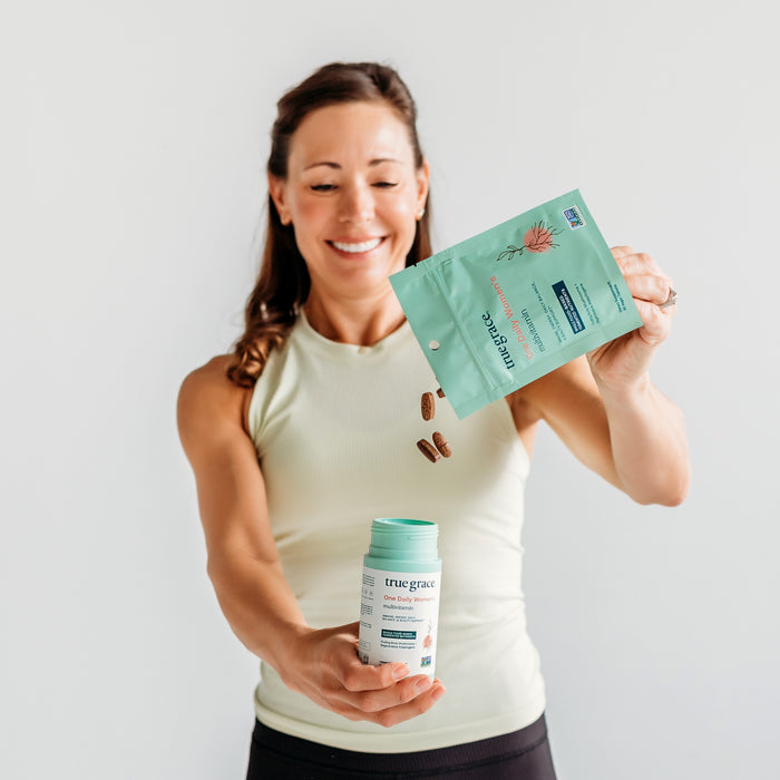 A woman in a tank top smiles while pouring capsules from a teal packet labeled One Daily Womens Multivitamin Refill by True Grace into a container, set against a plain, light background.