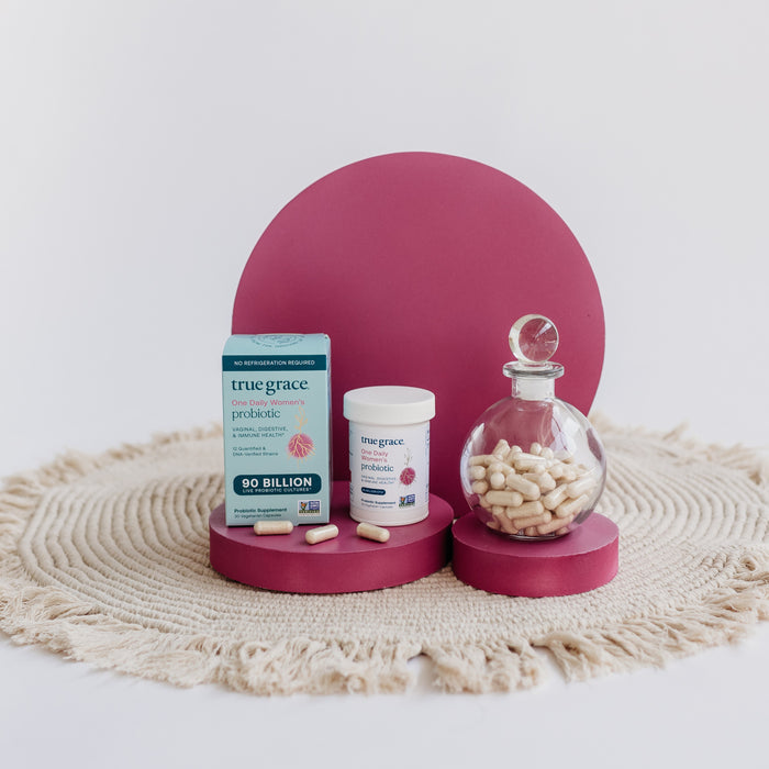 A textured beige mat features True Grace products for digestive health: a box and bottle of One Daily Womens Probiotic - Digestive, Vaginal & Immune Health, a glass jar, all on burgundy stands, with capsules scattered around.