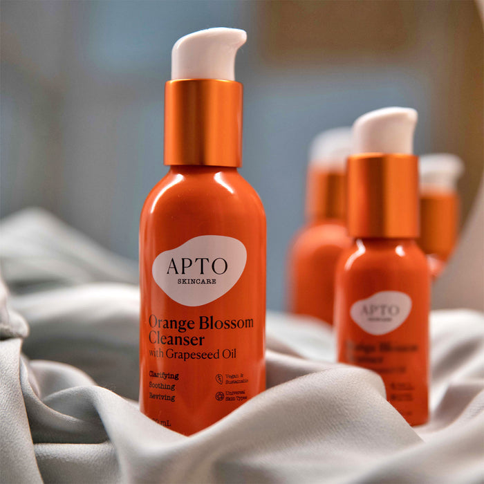 A cruelty-free APTO Skincare Orange Blossom Cleanser with Grapeseed Oil bottle featuring a white pump is showcased on silky fabric. Natural fruit ingredient bottles are blurred in the background, highlighting the orange color and its lightly foaming face wash formula.