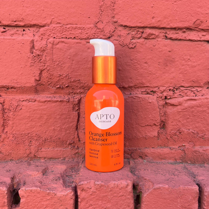 APTO Skincares bright orange bottle of Orange Blossom Cleanser with Grapeseed Oil, a lightly foaming face wash, is set against a textured red brick background. The cruelty-free product features a white pump and text detailing its size and benefits.