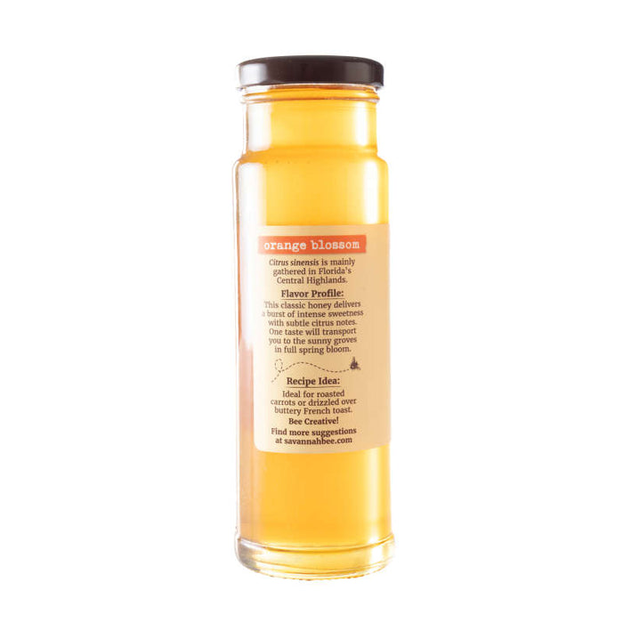 A tall Savannah Bee Company Orange Blossom Honey jar, rich with citrus aroma and golden-yellow hue, originates from sun-drenched orange groves. Perfect for drizzling on roasted carrots or ice cream, its label highlights the distinctive flavor profile.