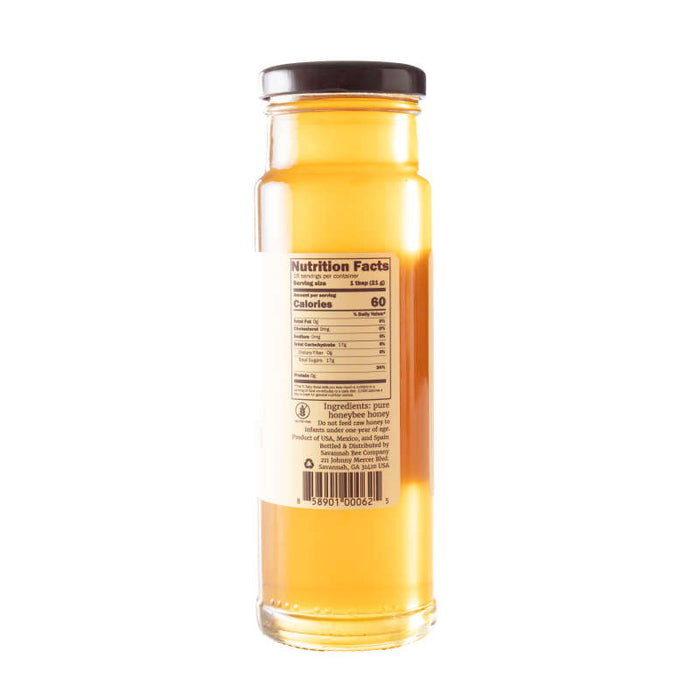 A tall jar of Savannah Bee Companys Orange Blossom Honey with a black lid has a label featuring nutrition facts, ingredients, and a barcode. The honey is golden with a subtle citrus aroma.