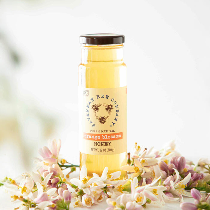 A glass jar of Savannah Bee Companys Pure & Natural Orange Blossom Honey, 12 oz (340 g), features a black lid and is encircled by flowers, sharing an enticing citrus aroma that evokes sunlit orange groves.