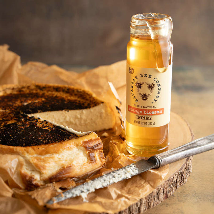 A jar of Savannah Bee Companys Orange Blossom Honey, exuding a citrus aroma, is beside toasted bread on parchment paper. A vintage-style knife with honey remnants lies in front, set against a rustic wooden surface.