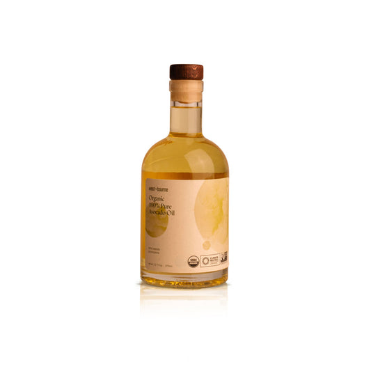 A clear glass bottle with a wooden cap, filled with yellow liquid, labeled "west~bourne Organic 100% Pure Avocado Oil" features chef-inspired graphics and certification logos on a beige label—ideal for any sustainable kitchen, isolated on white.