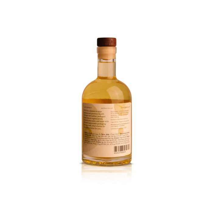 A clear glass bottle of west~bourne Organic 100% Pure Avocado Oil, filled with golden oil and sealed with a cork and wooden cap. A beige label with back text completes this eco-friendly kitchen essential, shown on a white background.