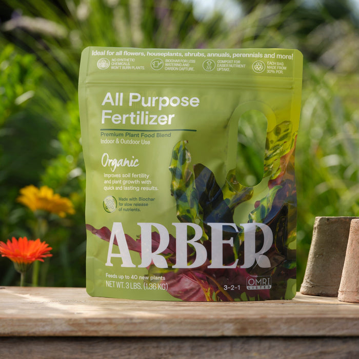 A green bag of Arbers Organic All Purpose Fertilizer with biochar is shown outdoors on wood. Its labeled organic for various plants, with bright greenery and an orange flower emphasizing its soil health benefits in the background.