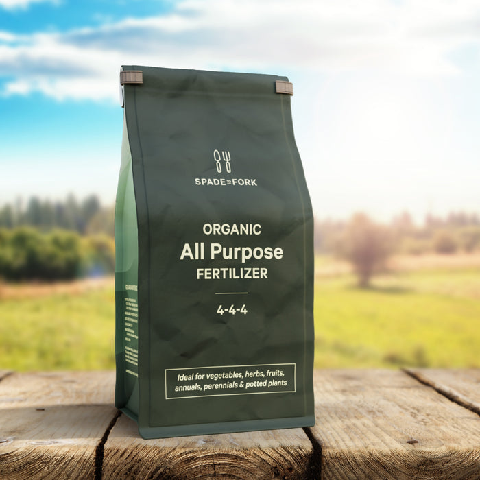 A green bag of Spade to Forks Organic All Purpose Fertilizer, containing a balanced 4-4-4 nutrient ratio, rests on wood. Its ideal for enhancing soil in vegetables, herbs, fruits, annuals, perennials, and potted plants. The backdrop features a scenic field.