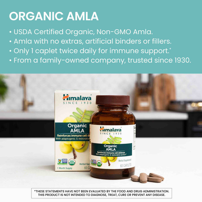 A bottle of Himalaya Organic Amla dietary supplement sits on a kitchen counter alongside its box. The label highlights its organic certification, non-GMO status, absence of artificial additives, and recommends taking one caplet twice daily for immune support.