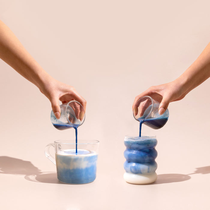 Two hands pour blume’s Organic Blue Lavender Latte Powder from small glasses into cups of milk, forming swirling blue patterns. The background is light beige, with one clear cylindrical cup and one wavy ceramic cup.
