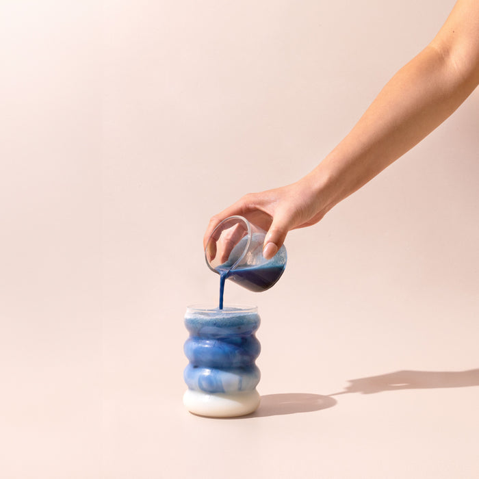 A hand pours deep blue Organic Blue Lavender Latte, made with blumes calming powder, from a glass into a wavy white-and-blue ceramic cup against a pale beige background.