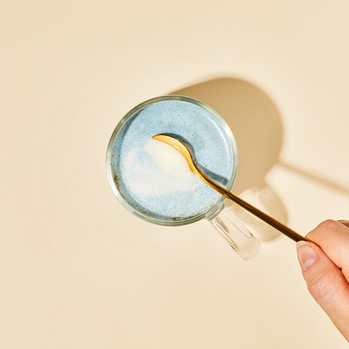 A hand stirs a cup of blume Organic Blue Lavender Latte Powder, creating a blue frothy bedtime drink with a gold spoon on a beige background, shadow visible.