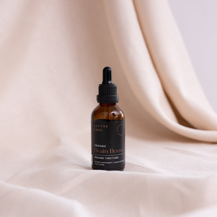 A small amber bottle labeled Organic Brain Boost Tincture by Sattva Soul rests on soft beige fabric, offering an elegant, minimalist look. Infused with Ayurvedic Brahmi, this neuro-protective elixir is crafted to support brain health.