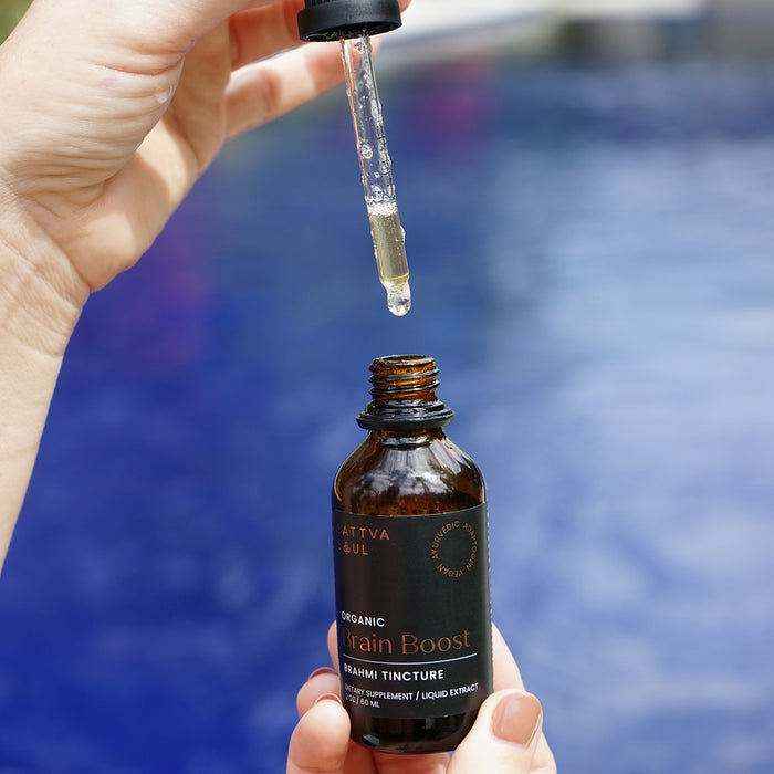 A hand holds a brown glass dropper bottle labeled Organic Brain Boost Tincture by Sattva Soul near a pool. Another hand releases drops of the neuro-protective elixir. A blurred blue water surface in the background enhances this tranquil Ayurvedic Brahmi ritual.