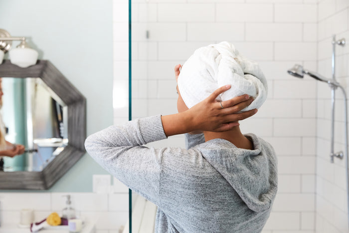 In a bathroom with white subway tiles, someone wearing a gray sweater adjusts their Grove Co. Organic Cotton Hair Towel wrapped around their head. The mirror reflects part of the room with toiletries on the sink.