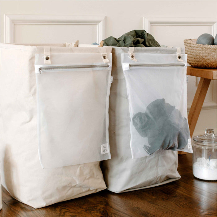 On a wooden floor, two Full Circle Organic Cotton Laundry Hampers with mesh pockets organize clothes—one holds green clothes and the other darks. A woven basket and glass jar of detergent enhance the aesthetic, blending functionality with sustainability.