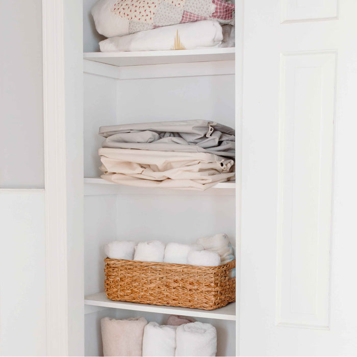 Open closet with neatly folded sheets and blankets on the shelves. A Full Circle Organic Cotton Laundry Hamper, filled with rolled white towels, sits on one shelf while additional towels are sorted below. The white walls enhance the clean and organized look of this sustainable setup.