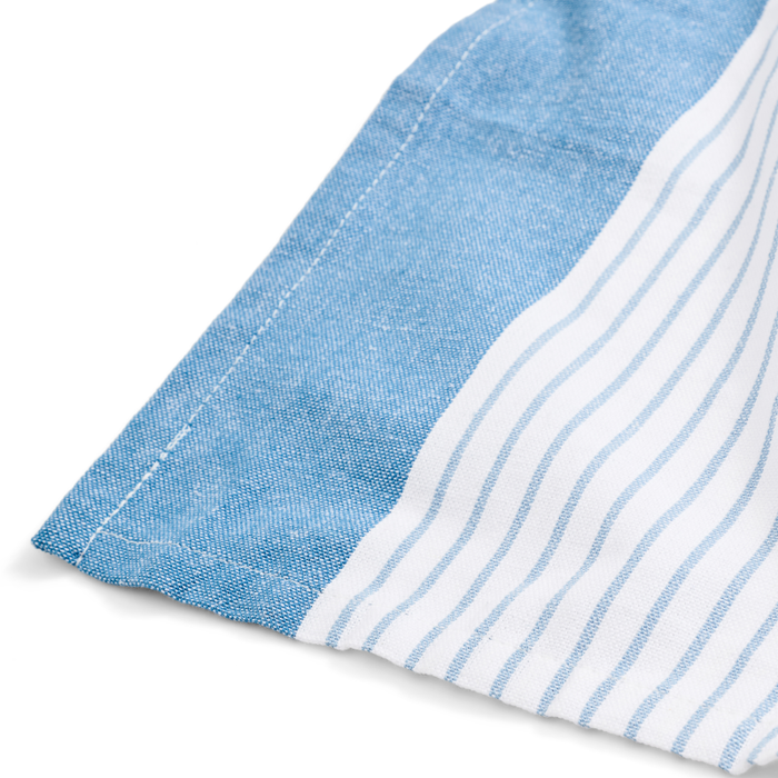 A close-up of Grove Co.s Organic Cotton Striped Kitchen Towels shows quick-drying blue chambray with visible stitching on the left and white fabric with thin blue vertical stripes on the right, connected at the seam. The chambray section is slightly folded.