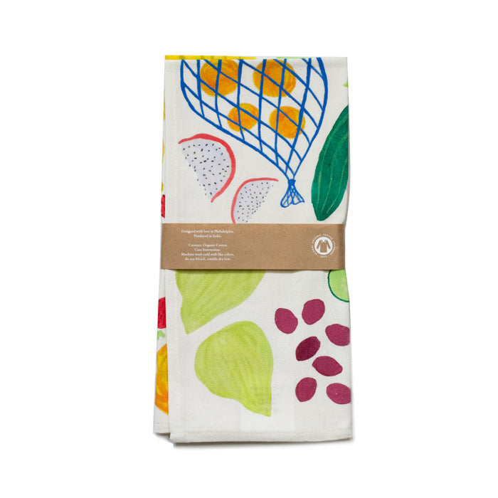 The Organic Cotton Tea Towel by Supra Endura is crafted from durable white organic cotton and features colorful fruit prints, including figs, leaves, and abstract shapes in yellow, blue, green, and red. It comes neatly folded with a brown paper band showcasing the original artwork.