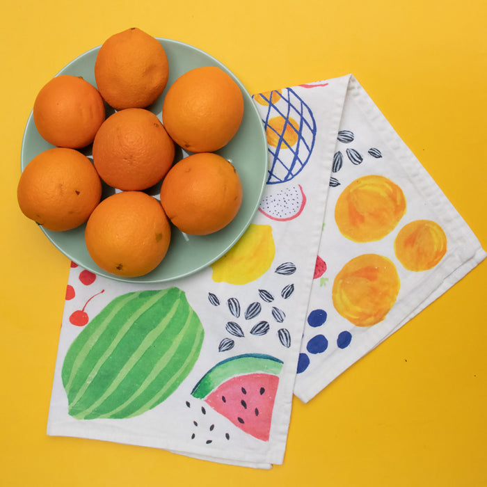 A green bowl of oranges sits on a vibrant yellow surface beside two Organic Cotton Tea Towels by Supra Endura, featuring durable organic cotton and adorned with colorful fruit designs including watermelon, cherries, and abstract patterns, showcasing original artwork.