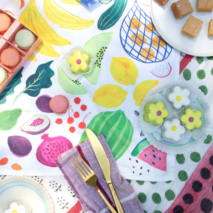 Top view of a colorful table setting with a vibrant fruit-patterned cloth, flower-shaped desserts, assorted macarons, caramel squares on a plate, and gold cutlery atop an original artwork-inspired Organic Cotton Tea Towel by Supra Endura.