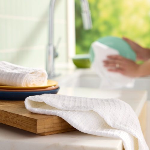 On a wooden board atop the kitchen counter sits a stack of Grove Co.s Organic Cotton Textured Kitchen Towels. In the background, someone holds a turquoise dish near the running sink. The bright, airy scene is enhanced by blurred greenery outside.