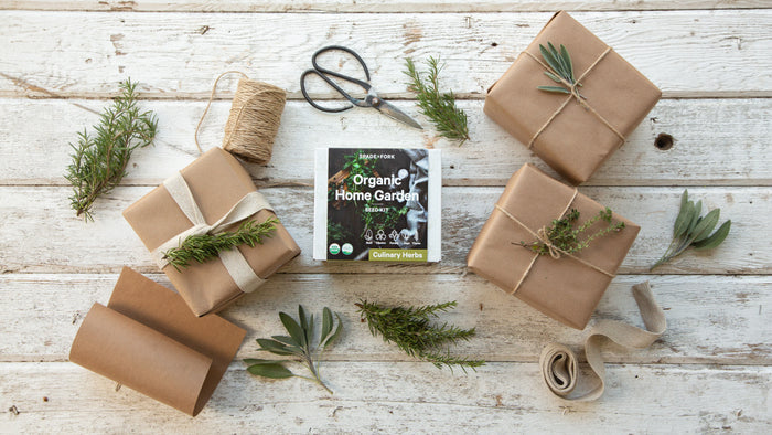 The Organic Culinary Herbs Kit by Spade to Fork is nestled among brown paper-wrapped boxes, twine, scissors, and herb sprigs on a rustic white wooden surface—ideal for creating your own oasis with organic seeds.