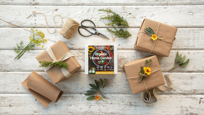 A top-down view shows four brown paper-wrapped gifts with flowers and leaves on a rustic table. In the center is Spade to Forks Organic Edible Flower Garden Kit, surrounded by twine, scissors, greenery sprigs, and culinary herb kits for your kitchen adventures.