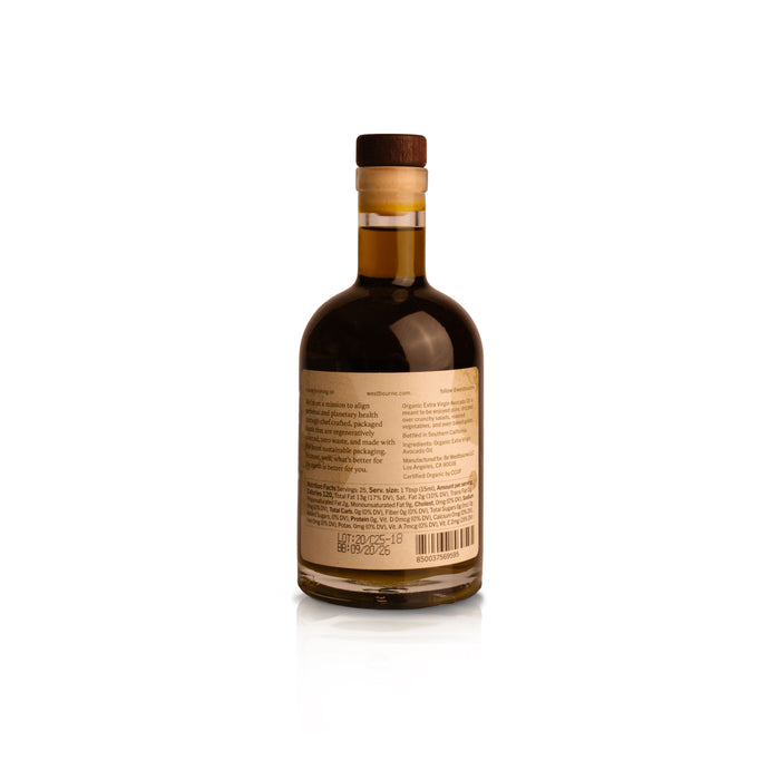A glass bottle of west~bourne Organic Extra Virgin Avocado Oil, filled with a dark liquid, stands upright on a white background. It features a wooden cap and beige label, ideal for high heat cooking, with text and a barcode on the back.