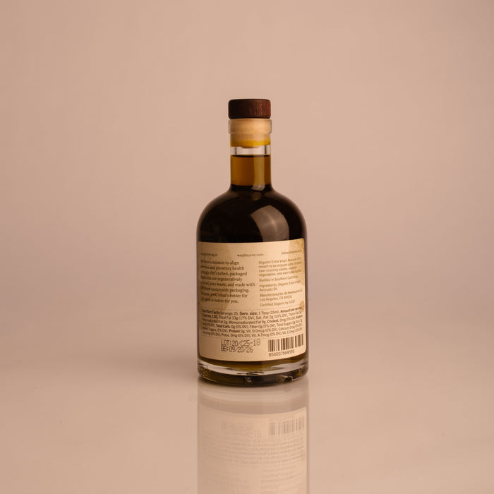 A small glass bottle of west~bourne Organic Extra Virgin Avocado Oil, filled with dark liquid and sealed with a wooden cap, features a barcode and label on the back—ideal for high-heat cooking or your sustainable kitchen, shown on a plain light background.