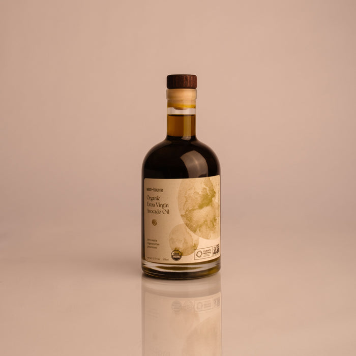 A glass bottle of west~bourne Organic Extra Virgin Avocado Oil with a wooden cap and abstract green label design—ideal for high heat cooking in sustainable kitchens, shown on a plain light background.