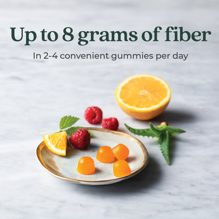 A plate with orange gummies, orange slices, raspberries, and green aloe leaves on a marble surface. Text above reads: "Up to 8 grams of fiber in 2-4 convenient gummies per day.