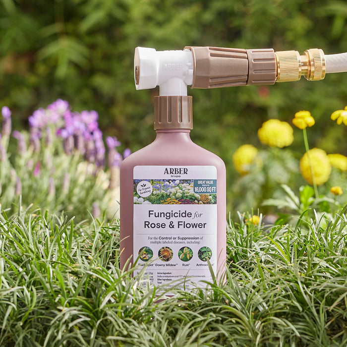 An Arber Organic Fungicide for Roses and Flowers, RTS bottle is attached to a garden hose among lush greenery and blooming flowers. Its label describes it as effective against plant diseases such as powdery mildew, with yellow and purple flowers visible in the background.