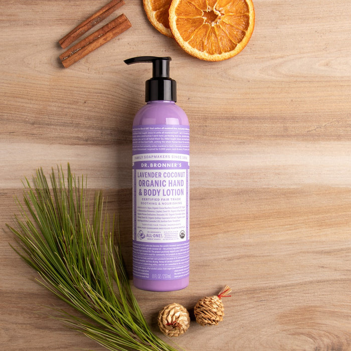 A bottle of Dr. Bronner’s Organic Hand & Body Lotion rests on a wooden surface surrounded by pine needles, pine cones, dried orange slices, and cinnamon sticks, emphasizing its soothing blend of organic oils.