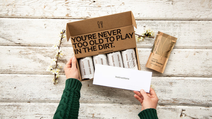 A person holds a box labeled YOURE NEVER TOO OLD TO PLAY IN THE DIRT, containing seed packets. Next to the box are booklets titled Instructions and Spade to Forks Organic Indoor & Outdoor Potting Soil Multi-Pack, all on a wooden surface.