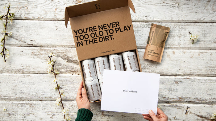 Someone holds an open cardboard box with plant seed packets and an instructional booklet on a rustic wooden surface sprinkled with white flowers. The lid reads, Youre never too old to play in the dirt, and contains a Spade to Fork Organic Indoor & Outdoor Potting Soil Multi-Pack.