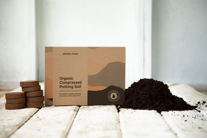 A box labeled Spade to Fork Organic Indoor & Outdoor Potting Soil Multi-Pack, featuring five discs rich in beneficial microorganisms, sits on a light wooden surface with a neutral background.