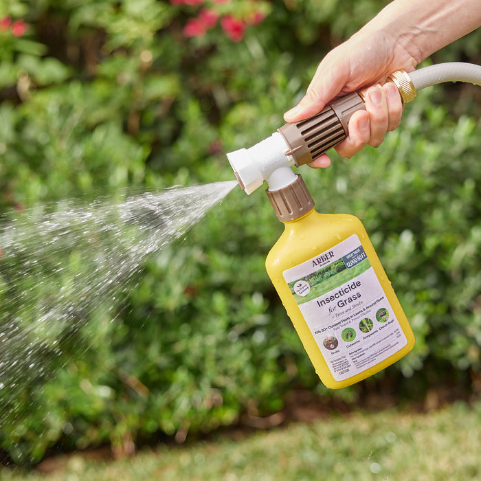 Using a hose attachment, someone sprays Arbers Organic Insecticide for Lawn, Tree, and Shrub RTS on the grass. The yellow bottle is labeled clearly for lawn pest control. A vibrant garden with lush green grass and pink flowers completes the background setting.