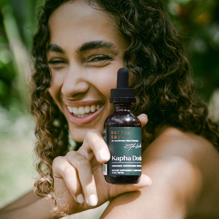 A smiling woman with curly hair holds an Organic Kapha Tincture - Energizing bottle by Sattva Soul, filled with Ayurvedic herb extracts. The background features blurred greenery, evoking a natural setting.