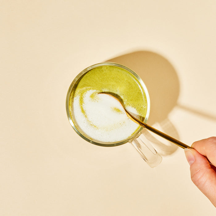A hand stirs a glass cup of blume Organic Matcha Coconut Latte Powder with a gold spoon, swirling the antioxidant-rich foam atop a beige background with shadow.