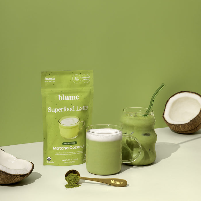 A package of blume Organic Matcha Coconut Latte Powder stands next to a glass mug of frothy green latte, an iced matcha with a straw, a halved coconut, and a gold spoon with vibrant matcha powder on a light green surface.