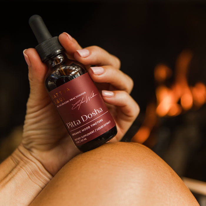 Someone holds a bottle of Sattva Souls Organic Pitta Tincture - Mood Support, ideal for those seeking organic balance. The label features gold and white text on maroon, with a warm firelight in the background evoking an Ayurvedic ambiance.