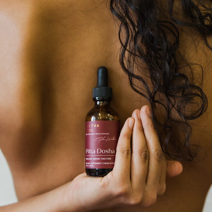A person with long, curly hair holds an amber bottle of Sattva Souls Organic Pitta Tincture - Mood Support against their chest. The bottle features a dropper and maroon labeling, while their skin and hair enhance the essence of an Ayurvedic practitioner.