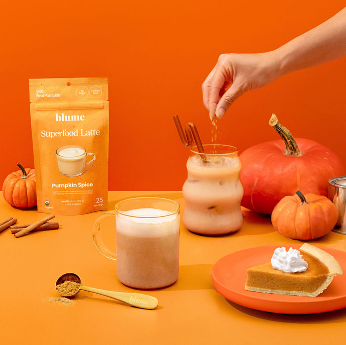 A hand sprinkles cinnamon into an iced latte. Nearby, theres an organic pumpkin, a packet labeled blume Organic Pumpkin Spice Latte Powder, a steaming mug, and pumpkin pie with whipped cream, highlighting the perfect blend of superfood ingredients.