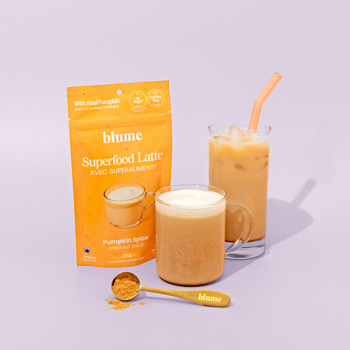 A pack of blumes Organic Pumpkin Spice Latte Powder is displayed beside an iced Pumpkin Spice Latte in a glass mug with a straw, next to a tall glass and a spoonful of the mix on a light purple surface.
