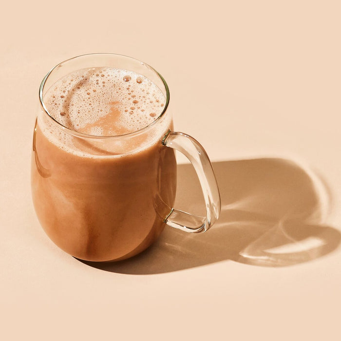 A brightly lit, minimalistic scene features a glass mug of blume Organic Reishi Hot Cacao Latte Powder, filled with frothy cocoa and casting a heart-shaped shadow on a light beige surface.