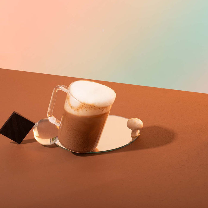 A glass mug of frothy coffee made with blume Organic Reishi Hot Cacao Latte Powder sits on a round mirror, beside a small mushroom, black geometric shape, and clear sphere on a brown surface with a softly colored gradient background.
